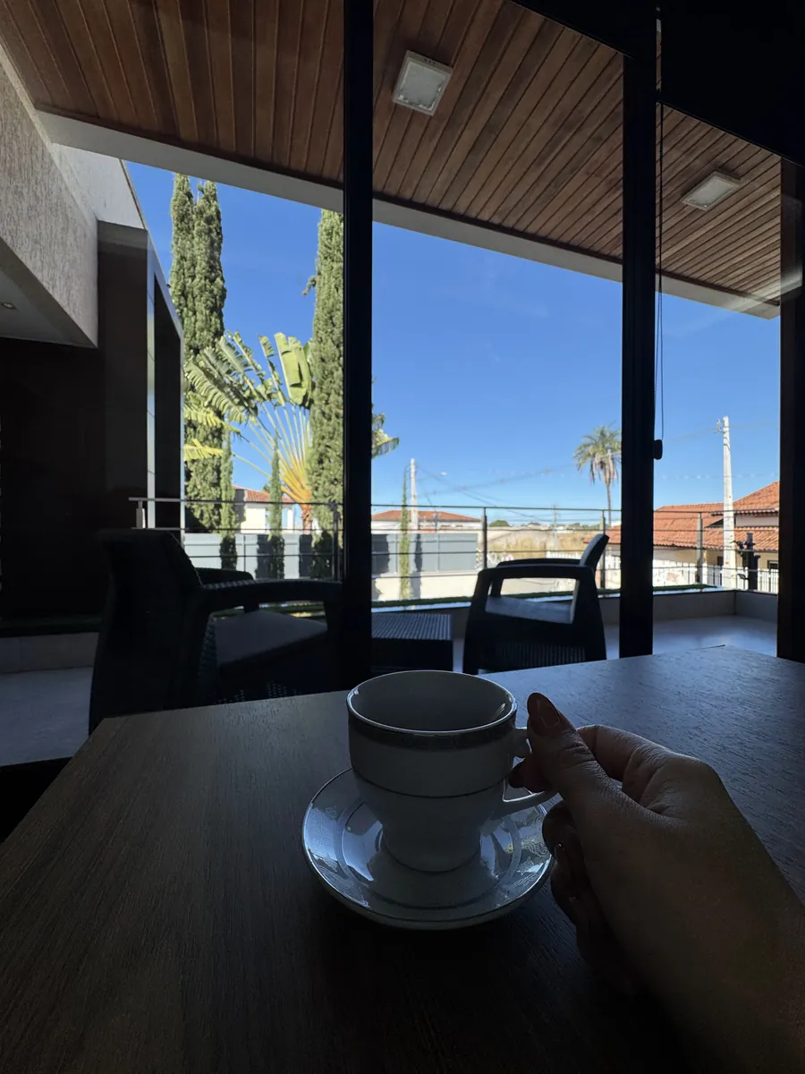 Mão segurando uma xícara de café sobre a mesa, com a varanda ao fundo