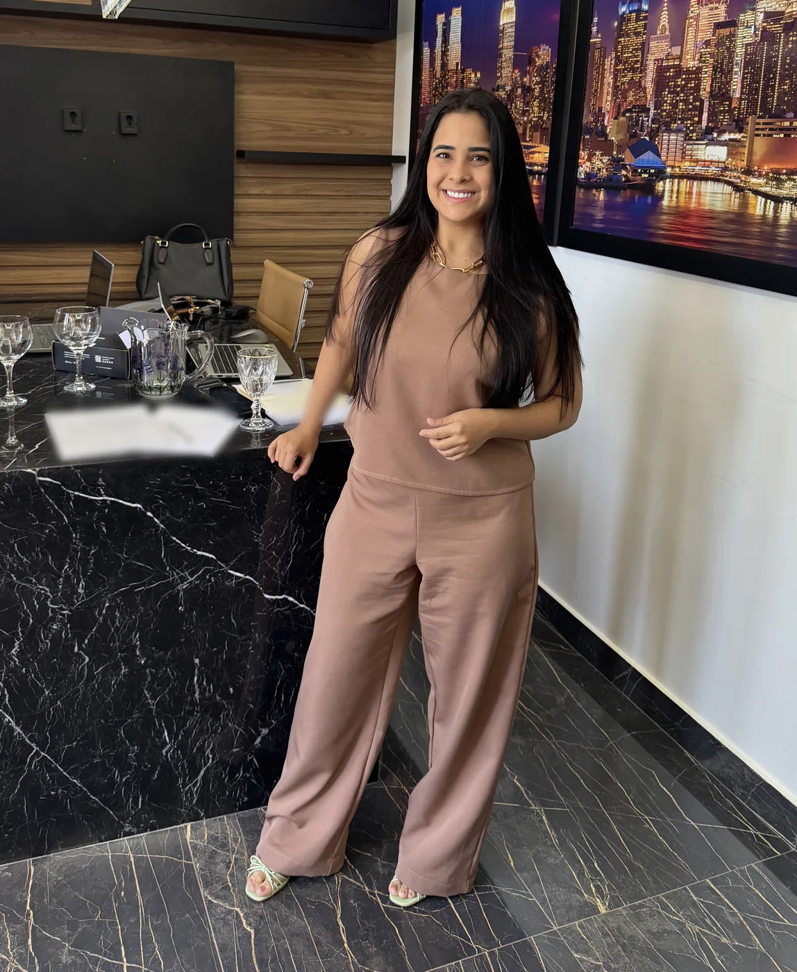Naila Jabour em pé junto à mesa de mármore negro do escritório, de conjunto rosé, sorrindo